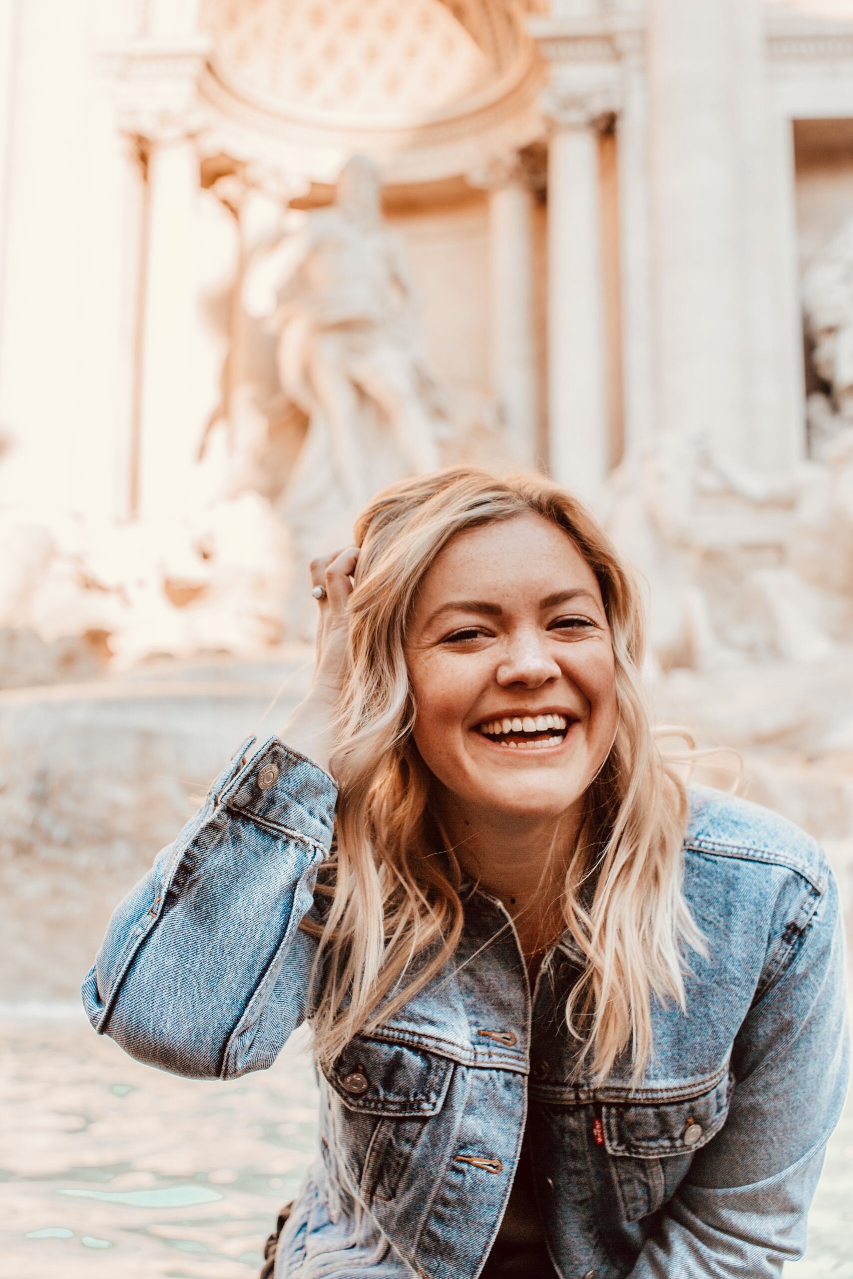 Veneers Neunkirchen - Dr. Alexander Puchner, MSc Symbolfoto Veneers: Breit lächelnde Frau vor einem Brunnen mit antiken Motiven auf den links oben helle Sonnenstrahlen fallen.